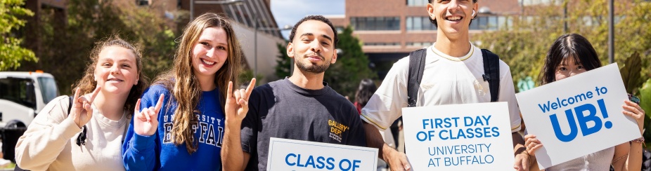 New UB students holding signs that say First day of classes and welcome to UB with North Campus in the background. 