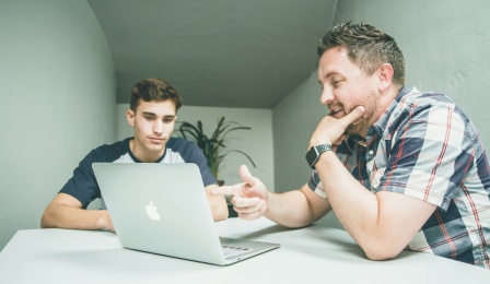 Two people sitting in front of a laptop, speaking. 