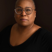 Photo shows a person in a black shirt against a black background, wearing glasses, looking into the camera. 
