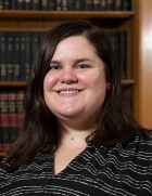 Image of a woman with shoulder-length brown hair wearing a black shirt with thin horizontal white stripes, smiling at the camera. A bookcase can be seen behind her. 