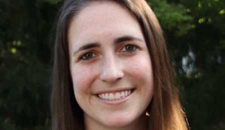 Photo of a person with long brown hair, smiling at the camera, outdoors. 