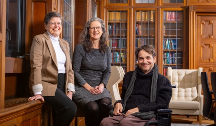 Photo of three people in a library, smiling at the camera. 