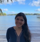 Photo of a woman - lake behind her, has long balck hair, wearing blue kurta , and smiling at the camera. 