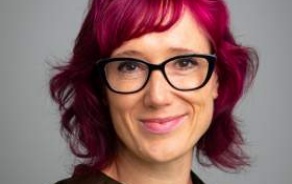 Photo shows a woman with pink hair and black glasses, smiling at the camera. 