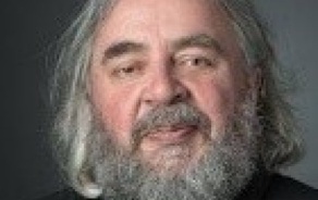 Photo shows a man with gray hair and a gray beard wearing a black shirt. 