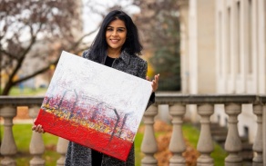 Photo shows a woman outdoors holding a painting. 