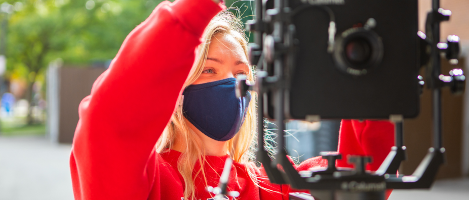 Photo shows a woman in a red shirt and mask using a camera to record an interview. 