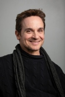 Man with short brown hair smiles at the camera wearing a dark sweater and matching scarf. 
