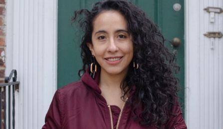 Woman standing in front of a green door, wearing a purple shirt, smiling at the camera. 
