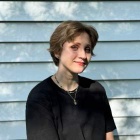 Image of Deirdre Harder, who has short, brown hair, wearing a black T-Shirt, in the shadow of a tree on a sunny day. 