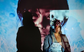 Chanee Choi's Remembrance: Mapping Memory Through Bio-Interactive Media. This image contains the artist, wearing a headset which collects data, standing in front of a blue projection, which depicts a close-up of a woman. 