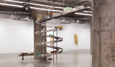 Telford Keough's Carriers (Gravity-Fed), (2024) is a dynamic sculptural installation that contains mold-blown glass vessels carrying a combination of organic matter and industrial waste, spaced apart on a static sprial conveyor belt; made of stainless steel, stained glass, commercial signage, Gowanus Canal Sediment, soap, and various found domestic items. 