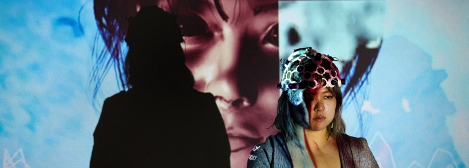 Chanee Choi's Remembrance: Mapping Memory Through Bio-Interactive Media. This image contains the artist, wearing a headset which collects data, standing in front of a blue projection, which depicts a close-up of a woman. 