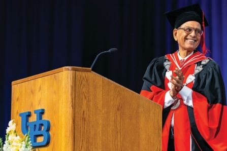 UB President Satish Tripathi in academic regalia at a podium clapping for graduates in 2023. 