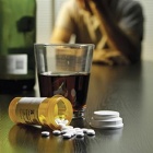 A bottle and glass of alcohol next to a spilled bottle of pills with a man in the background. 