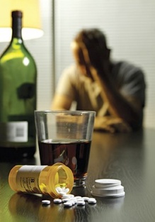 A bottle and glass of alcohol next to a spilled bottle of pills with a man in the background. 