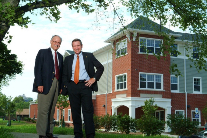 Two men in suits standing in front of a brick and shingled building. 