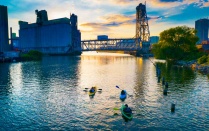 People in kayaks paddle the in the Buffalo River with grain elevators in the distance. 