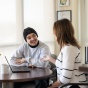 two people talking and sitting at a table with a laptop and small plant. 