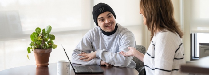 two people talking and sitting at a table with a laptop and small plant. 