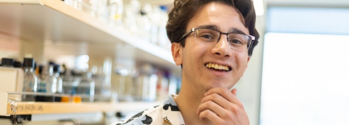 student smiling in a lab with rows of lab equipment in background. 