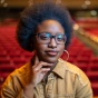 person with slight smile posing in theatre with chairs in background. 