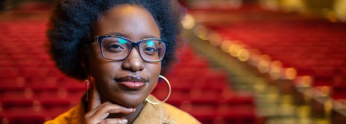 person with slight smile posing in theatre with chairs in background. 