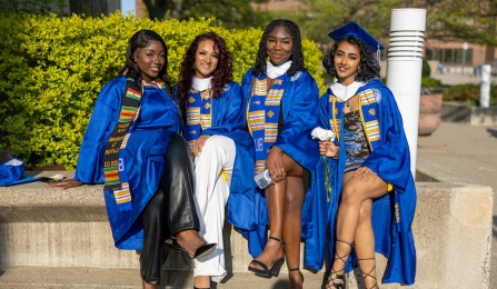 four students sitting on a concrete ledge wearing graduation caps and gowns. 
