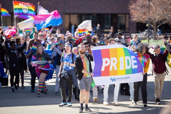 UB community members marching down the academic spine on North Campus with raibbow flags and a banner that reads "PRIDE" with rainbow colors. 