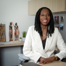 Anyango Kamino smiling in a white blazer and black shirt sitting at a desk. 