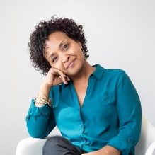 Rahwa Ghirmatzion smiling in a green blouse sitting in a white chair on a white background. 