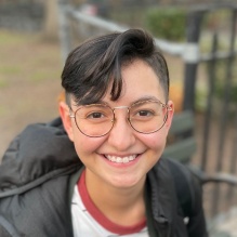 Lina Tabak smiling in a black jacket and white and red shirt sitting on a bench background. 