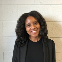 Arturia Melson-Silimon smiling in a black blazer and black shirt on a wall background. 