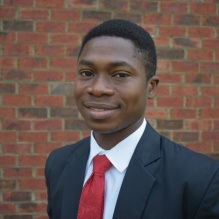 Babatunde Keshinro smiling in a headshot with a suit and tie in outside background with bricks in the back. 