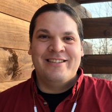 Waylon Wilson smiling with red shirt and wood house in the background. 