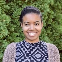 Yemimah King smiling with patterned shirt and swaeater in outdoor background with trees. 