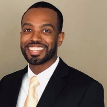 Joshua Cloudy smiling in a headshot with a dark suit, a tie and white shirt in a indoor clear background. 