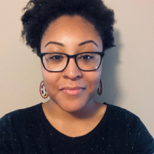 Erica Banks wearing reading glasses and smiling infront of a solid backdrop. 