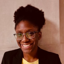 AraOluwa Adaramola smiling in a yellow shirt and black blazer on a stone wall background. 