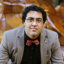 Christian Brickhouse smiling in a black shirt, red bowtie, and gray blazer and sitting on a mosaic chair. 