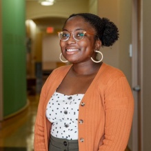 Julia Brisbane smiling in a white shirt with polka dots and an orange cardigan on a hallway background. 
