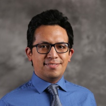 Jose Capa Salinas smiling in a blue shirt and gray tie on a gray background. 