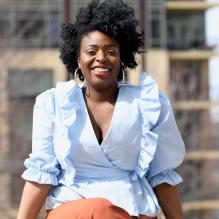 Khrysta Evans smiling in a blue shirt while sitting in front of a building. 