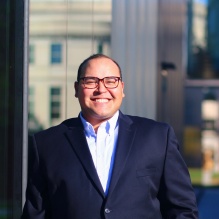 Efraín Rodríguez-Ocasio smiling in a black blazer and white shirt on an outside background. 