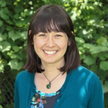Katherine Lebioda smiling in a black shirt with multicolored flowers, green cardigan, and green necklace on a background of a fence with leaves on it. 
