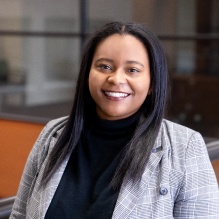 Khadejah Ray smiling in a gray blazer and black turtleneck sweater on a window background. 