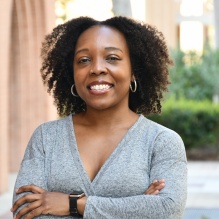 Meki Singleton smiling in a gray sweater on an outside background. 
