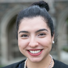 Elizabeth Barahona smiling in a headshot with a dark shirt and sweater with a pearl necklace in outside background with brick building. 