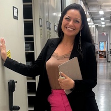 Jessica Fremland smiling in a brown shirt, pink pants, and black blazer on a hallway background. 