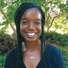 Tosin Adesogan smiling in a black shirt and a necklace saying her name on a background of trees. 